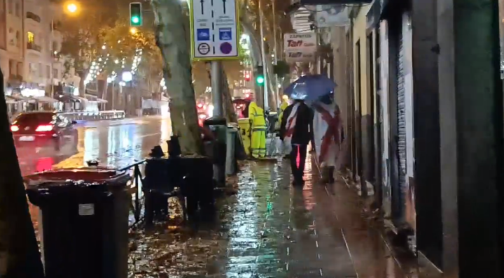 Dos manifestantes con banderas con cruces de Borgoña se refugian bajo un paraguas en la calle del Marqués de Urquijo mientras los empleados municipales limpian la calle.